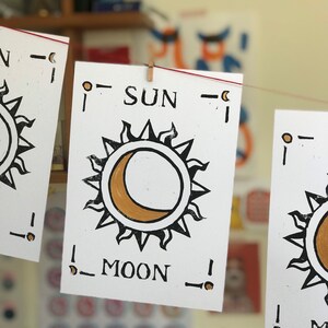 May include: White cards with black and gold sun and moon illustrations are strung on a red line. The word "SUN" is printed above the sun graphic, and "MOON" is below the moon graphic. The cards are held by small wooden pegs.