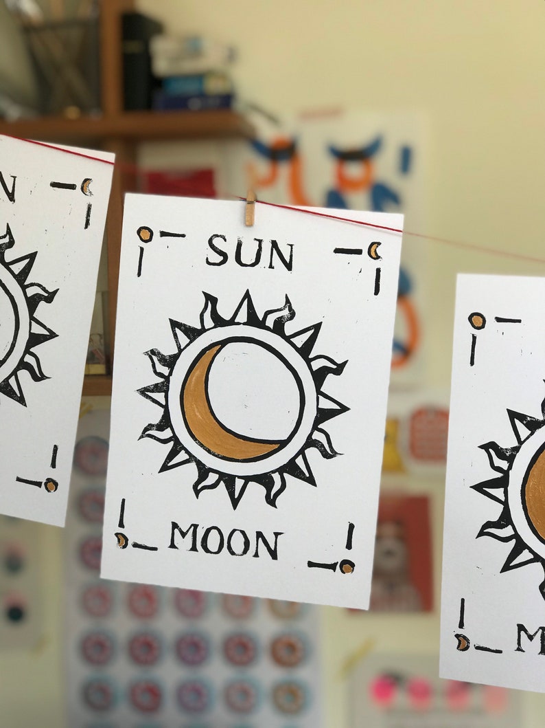 May include: White cards with black and gold sun and moon illustrations are strung on a red line. The word "SUN" is printed above the sun graphic, and "MOON" is below the moon graphic. The cards are held by small wooden pegs.