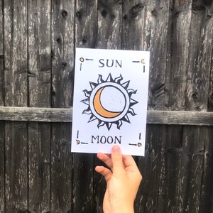 May include: A white card with the words "SUN" and "MOON" printed in black. The card features a sun and moon design with orange and black accents. The card is held in front of a weathered wooden fence.