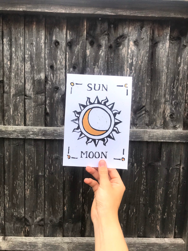 May include: A white card with the words "SUN" and "MOON" printed in black. The card features a sun and moon design with orange and black accents. The card is held in front of a weathered wooden fence.