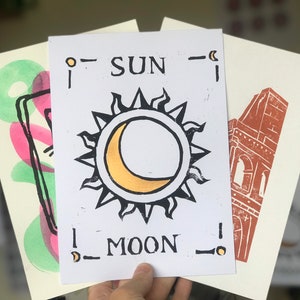 May include: A collection of art prints with linocut designs. The central print shows a sun and moon with the words "SUN" and "MOON". Other prints feature abstract shapes and a building.
