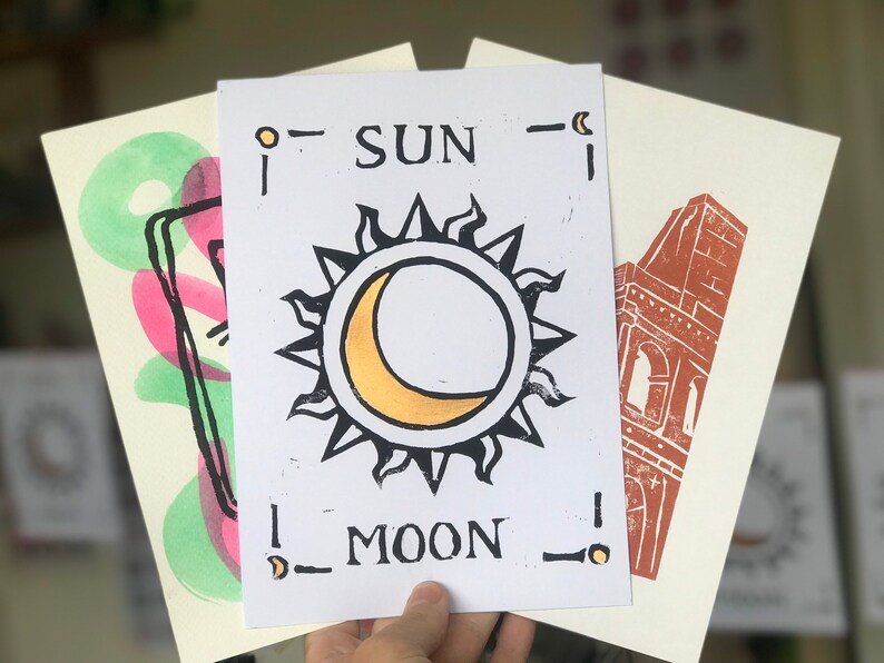 May include: A collection of art prints with linocut designs. The central print shows a sun and moon with the words "SUN" and "MOON". Other prints feature abstract shapes and a building.