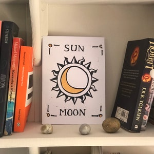 May include: A white card with a sun and moon design, with the words "SUN" and "MOON" printed on it, is displayed on a shelf. The card is flanked by books and small decorative stones. The sun is yellow and the moon is a yellow crescent.
