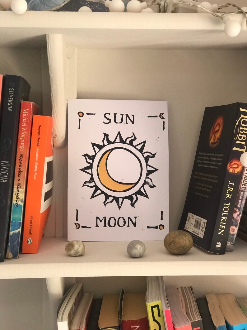 May include: A white card with a sun and moon design, with the words "SUN" and "MOON" printed on it, is displayed on a shelf. The card is flanked by books and small decorative stones. The sun is yellow and the moon is a yellow crescent.