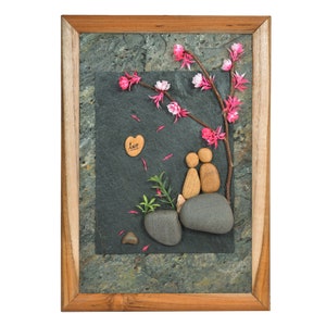May include: A framed artwork featuring a couple made of stones sitting under a pink blossom tree. The artwork is on a slate background and includes a small heart with the word "love" written on it.
