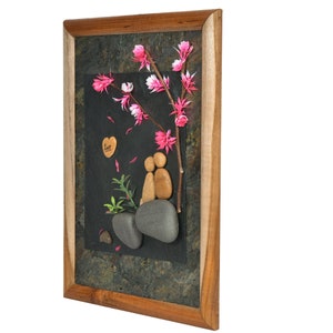 May include: A framed artwork featuring a couple made of stones, sitting under a branch with pink blossoms. The background is a dark slate with the word "love" written on a heart-shaped stone.