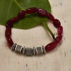 May include: A red gemstone bracelet with silver square beads. The bracelet is made of oval-shaped red stones and has four silver square beads with a textured surface.