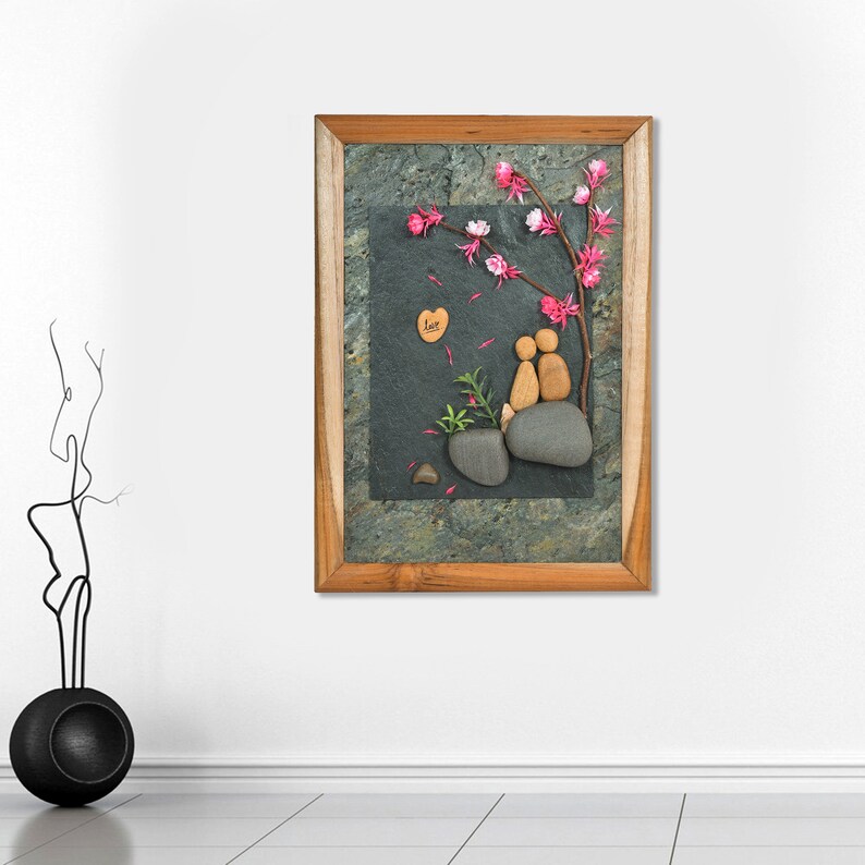 May include: A framed artwork featuring a couple made of stones sitting under a pink blossom tree. The artwork is on a slate background with a wooden frame. The word "love" is written on a small heart-shaped piece of wood.
