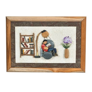 May include: A framed artwork made of small stones depicting a person sitting in a chair reading a book, a bookshelf, a lamp, and a flower in a vase.