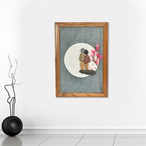 May include: A framed pebble art piece depicting two figures standing on a wooden base, set against a white circle and gray background. Pink blossoms adorn the right side. The frame is made of wood.