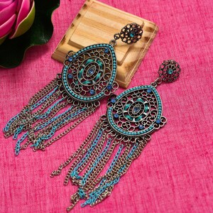 May include: A pair of ornate, teardrop-shaped earrings with intricate filigree detailing. The earrings feature turquoise and blue accents, including small beads and chain fringe. The metal has a bronze finish, and the earrings are displayed on a pink surface.