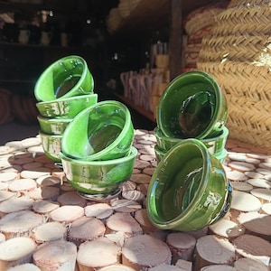 Puede incluir: Una colección de pequeños cuencos de cerámica verde brillante. Los cuencos están apilados y colocados sobre una superficie de madera. Los cuencos tienen forma redondeada con un borde ligeramente ensanchado. Tema: menaje de cocina.