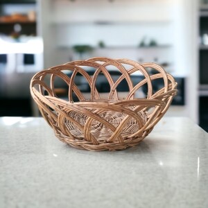May include: A woven wicker basket with a rounded shape. The basket is made of light brown wicker and has an open weave design. The basket is sitting on a light-colored countertop.