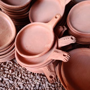 May include: A collection of terracotta clay pans with round bowls and short handles. Each handle features a small hole at the end and decorative etched zigzag patterns. The pans are stacked in multiple piles, showcasing the natural reddish-brown color.