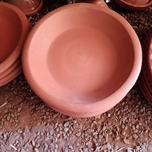 Può includere: Una ciotola in argilla color terracotta con una superficie interna liscia e ampia e un bordo spesso e arrotondato. La ciotola presenta una finitura opaca e una tonalità terrosa coerente, mostrando un design semplice e funzionale.