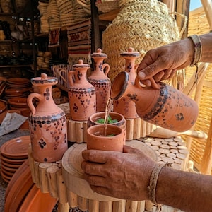 May include: A collection of terracotta pottery, including pitchers and cups, displayed on a wooden stand. The pitchers have handles and lids, decorated with dark brown floral patterns. Water is being poured from a pitcher into a cup with mint leaves.