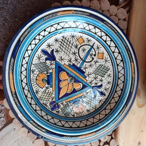 Puede incluir: Un plato decorativo de cerámica con un diseño geométrico azul, blanco y naranja. El plato presenta un diseño central con motivos florales y geométricos, rodeado de bandas concéntricas azules y blancas. El plato tiene aproximadamente 30 cm de diámetro.