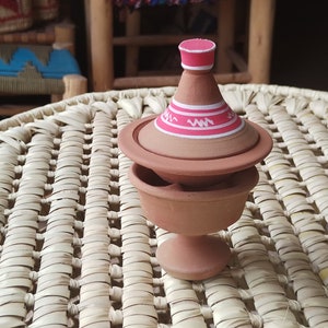 Op de afbeelding: Een terracotta-kleurig keramisch vat met deksel en voet. Het deksel heeft een roze band met witte zigzagontwerpen. Het vat staat op een geweven, lichtgekleurde mat, wat wijst op een cultureel of decoratief item.
