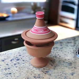 May include: A terracotta tagine pot with a conical lid, featuring pink and white painted details. The pot has a round base and a pedestal. The lid has a pink top and white zig-zag design. The tagine is designed for cooking.