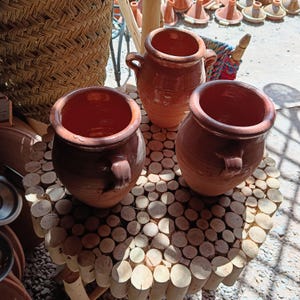 May include: Three terracotta pots with handles sit on a round wooden table made of small, cut logs. The pots are various sizes and have a reddish-brown colour. A woven basket is visible in the background, along with other pottery.