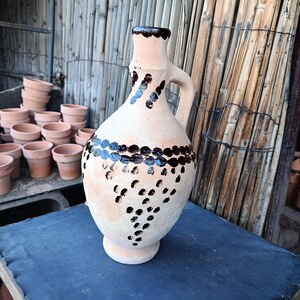 Puede incluir: Una jarra de cerámica beige con borde y asa marrón oscuro. La jarra está decorada con una banda de puntos marrones oscuros y puntos más pequeños dispersos por el cuerpo. Una pila de pequeñas macetas de terracota es visible en el fondo.