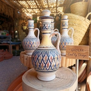 Op de afbeelding: Een verzameling decoratieve keramische kruiken met ingewikkelde patronen in blauw, geel en rood. Elke kruik heeft een handvat en een deksel. De kruiken worden tentoongesteld op houten standaards. Het bord luidt "GHOLFA - Cool by Design."