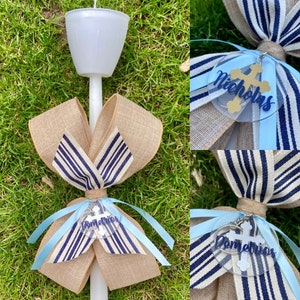 May include: A white candle with a burlap bow featuring blue and white striped ribbon. A light blue ribbon and a clear charm with a gold cross and the name "Demetrios" are attached. The candle is on a green grassy surface.