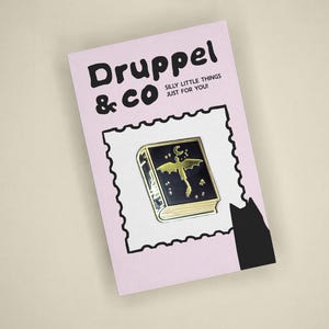 May include: A rectangular light pink card featuring the text Druppel & CO SILLY LITTLE THINGS JUST FOR YOU! and a black silhouette of a cat. Centered is a black enamel pin shaped like a book with a gold dragon, moon, and stars design.
