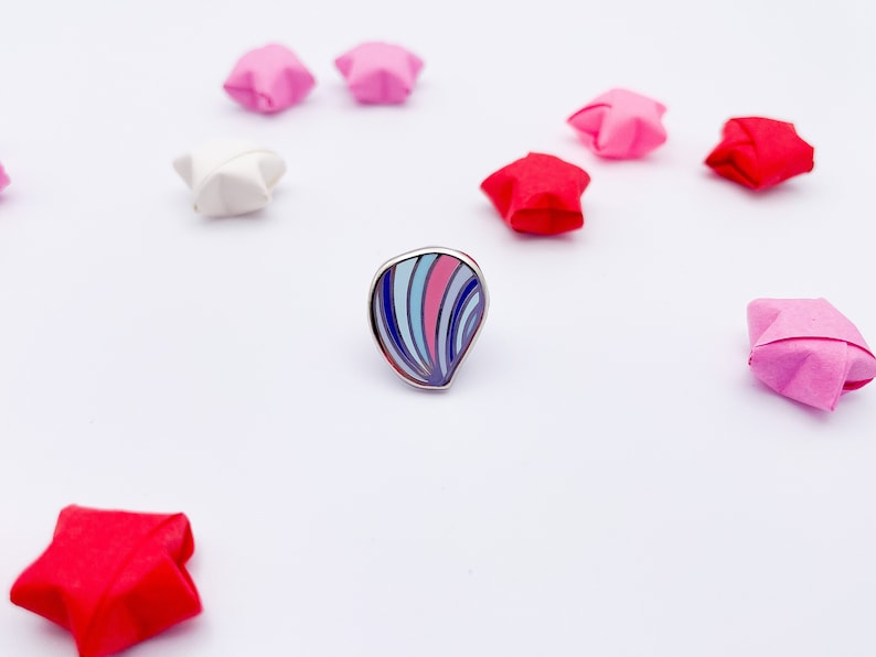 May include: A teardrop-shaped enamel pin featuring a silver metal border and a pattern of curved stripes in shades of dark blue, light blue, and pink. The pin is surrounded by several small, folded paper stars in red, pink, and white colours.