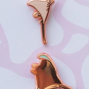 May include: Two rose gold metal enamel pins shaped like stingrays. The top pin features a light beige enamel fill, while the bottom pin has a polished rose gold finish with a small teal accent on the upper edge and tiny white dots at the base.