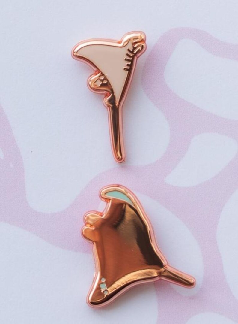 May include: Two rose gold metal enamel pins shaped like stingrays. The top pin features a light beige enamel fill, while the bottom pin has a polished rose gold finish with a small teal accent on the upper edge and tiny white dots at the base.
