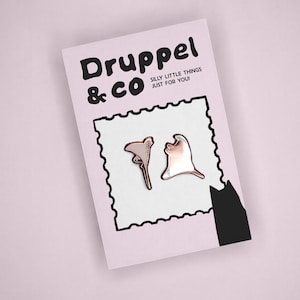 May include: Two metallic rose gold pins shaped like abstract organic forms displayed on a light pink backing card. The card features the text Druppel & CO, the phrase SILLY LITTLE THINGS JUST FOR YOU!, a black stamp-like border, and a black cat silhouette.
