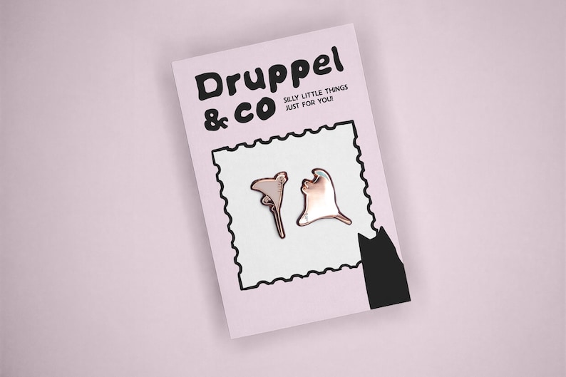 May include: Two metallic rose gold pins shaped like abstract organic forms displayed on a light pink backing card. The card features the text Druppel & CO, the phrase SILLY LITTLE THINGS JUST FOR YOU!, a black stamp-like border, and a black cat silhouette.