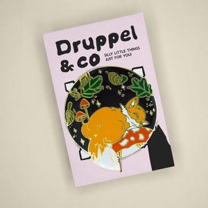 May include: A round enamel pin featuring an orange fox sleeping on a red and white mushroom against a black background with green leaves and small stars. The pin is attached to a light pink backing card with the text Druppel & CO SILLY LITTLE THINGS JUST FOR YOU!