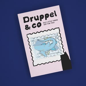 May include: A light pink rectangular card featuring the black text Druppel & CO SILLY LITTLE THINGS JUST FOR YOU! and a central illustration of a blue shark inside a white postage stamp border with a black silhouette shape at the bottom right.