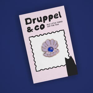 May include: A light purple enamel pin shaped like an open clam shell with a dark blue pearl in the center, attached to a light pink backing card with the text Druppel & CO SILLY LITTLE THINGS JUST FOR YOU and a black cat silhouette.