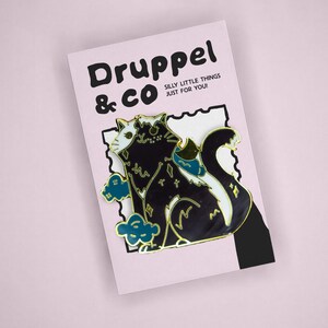 May include: A black and white cat enamel pin with gold-tone outlines, featuring small star accents and blue cloud shapes. The pin is attached to a light pink backing card with the text Druppel & CO SILLY LITTLE THINGS JUST FOR YOU printed on it.