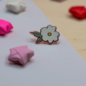 May include: A white flower enamel pin with a gold outline and a yellow centre, with origami stars in pink, red, and white. The pin has a gold-coloured stem and green leaves.