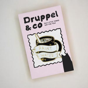 May include: A rectangular light pink backing card with the text Druppel & CO SILLY LITTLE THINGS JUST FOR YOU! featuring a black enamel pin of a mug with a white banner reading PAINT WATER and a black cat silhouette in the bottom right corner.