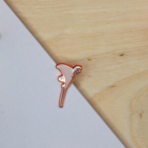 May include: A rose gold metal enamel pin shaped like a martini glass with a light pink fill. The glass features a small decorative detail on the side and the text "WLS" engraved in a stylized font along the upper edge of the triangular bowl.