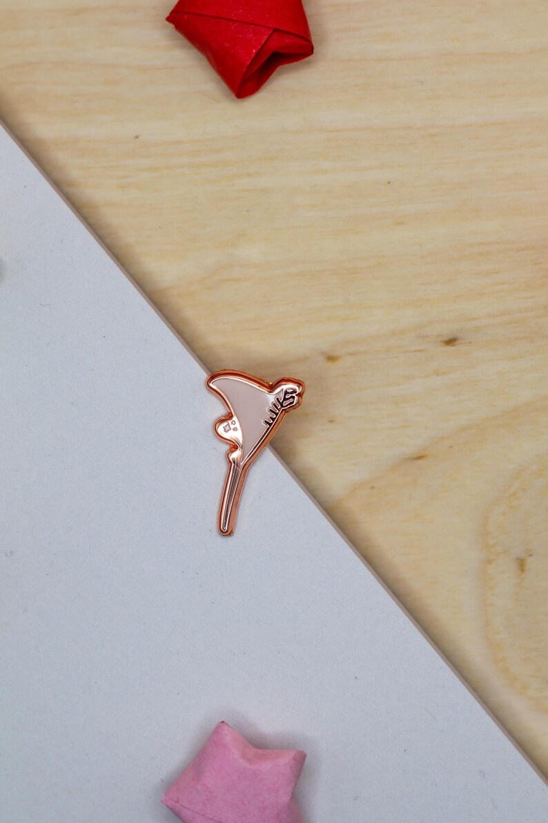 May include: A rose gold metal enamel pin shaped like a martini glass with a light pink fill. The glass features a small decorative detail on the side and the text "WLS" engraved in a stylized font along the upper edge of the triangular bowl.