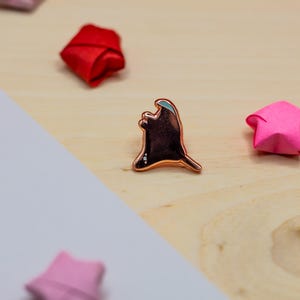 May include: A copper-toned metal enamel pin shaped like a ghost with a smooth, dark brown center and a metallic outline. The pin is surrounded by several small, folded paper stars in shades of red, pink, and white on a light wooden surface.