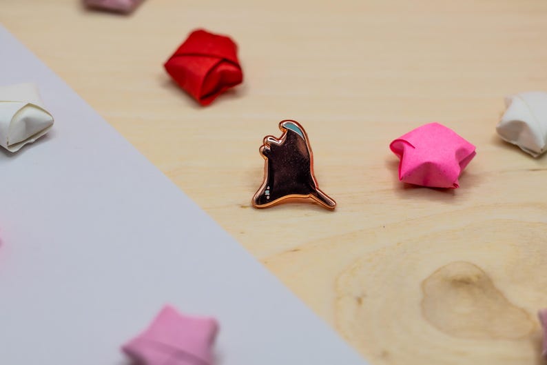 May include: A copper-toned metal enamel pin shaped like a ghost with a smooth, dark brown center and a metallic outline. The pin is surrounded by several small, folded paper stars in shades of red, pink, and white on a light wooden surface.