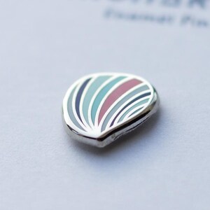 May include: A small, teardrop-shaped enamel pin featuring a series of curved, radiating stripes in shades of light blue, dark blue, and muted red. The design is encased in a polished silver-toned metal border, creating a smooth, rounded edge.
