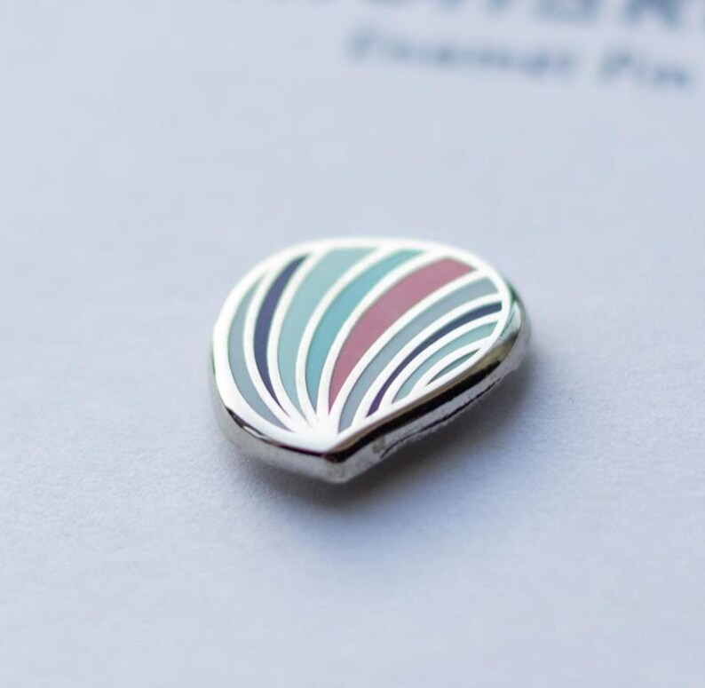 May include: A small, teardrop-shaped enamel pin featuring a series of curved, radiating stripes in shades of light blue, dark blue, and muted red. The design is encased in a polished silver-toned metal border, creating a smooth, rounded edge.