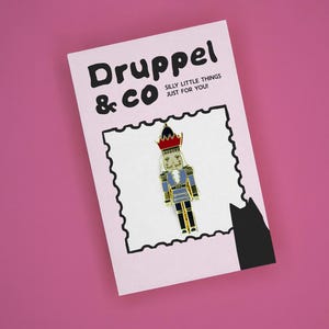 May include: A metal nutcracker enamel pin featuring a red hat, blue jacket, and black trousers, attached to a light pink backing card with a black scalloped border. The card displays the text Druppel & CO SILLY LITTLE THINGS JUST FOR YOU!