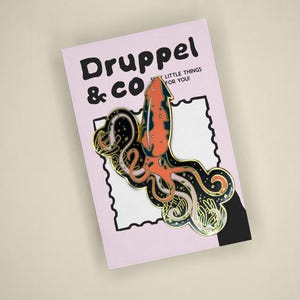 May include: A metal enamel pin featuring a stylized orange and black squid with long tentacles, displayed on a light pink backing card with the text Druppel & CO and the phrase SILLY LITTLE THINGS FOR YOU printed in black ink at the top.