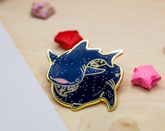 May include: A dark blue enamel pin shaped like a whale shark with a gold metal outline. The body of the shark features a white speckled pattern resembling a starry night sky, with a small pink mouth and gold accents detailing the fins and gills.