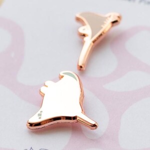 May include: Two rose gold metal pins shaped like stylized ghosts with smooth, reflective surfaces. Each pin features a rounded head and a pointed tail, showcasing a metallic finish that catches the light. The pins are displayed on a light-colored backing.