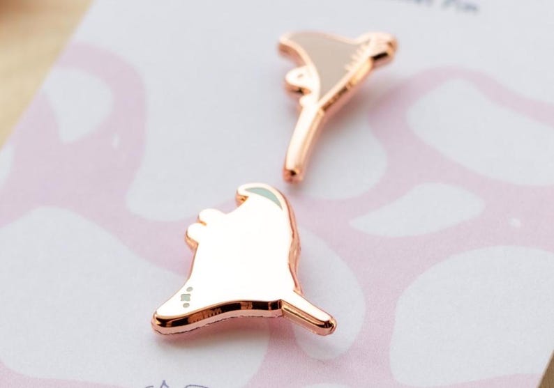 May include: Two rose gold metal pins shaped like stylized ghosts with smooth, reflective surfaces. Each pin features a rounded head and a pointed tail, showcasing a metallic finish that catches the light. The pins are displayed on a light-colored backing.